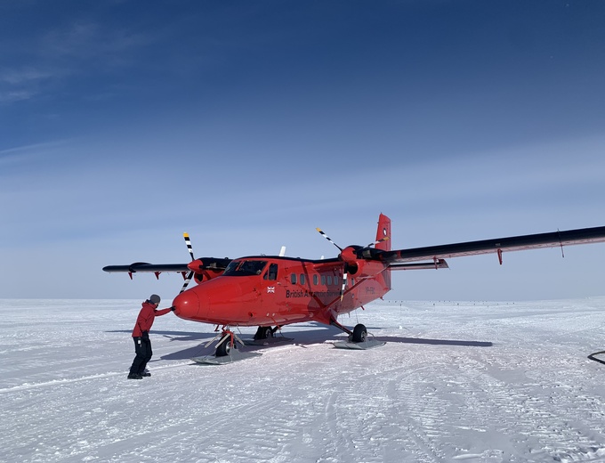 Aa powered down small red twin propeller aircraft sits on a flat field of snow. A man in a red jacket is poking the front 